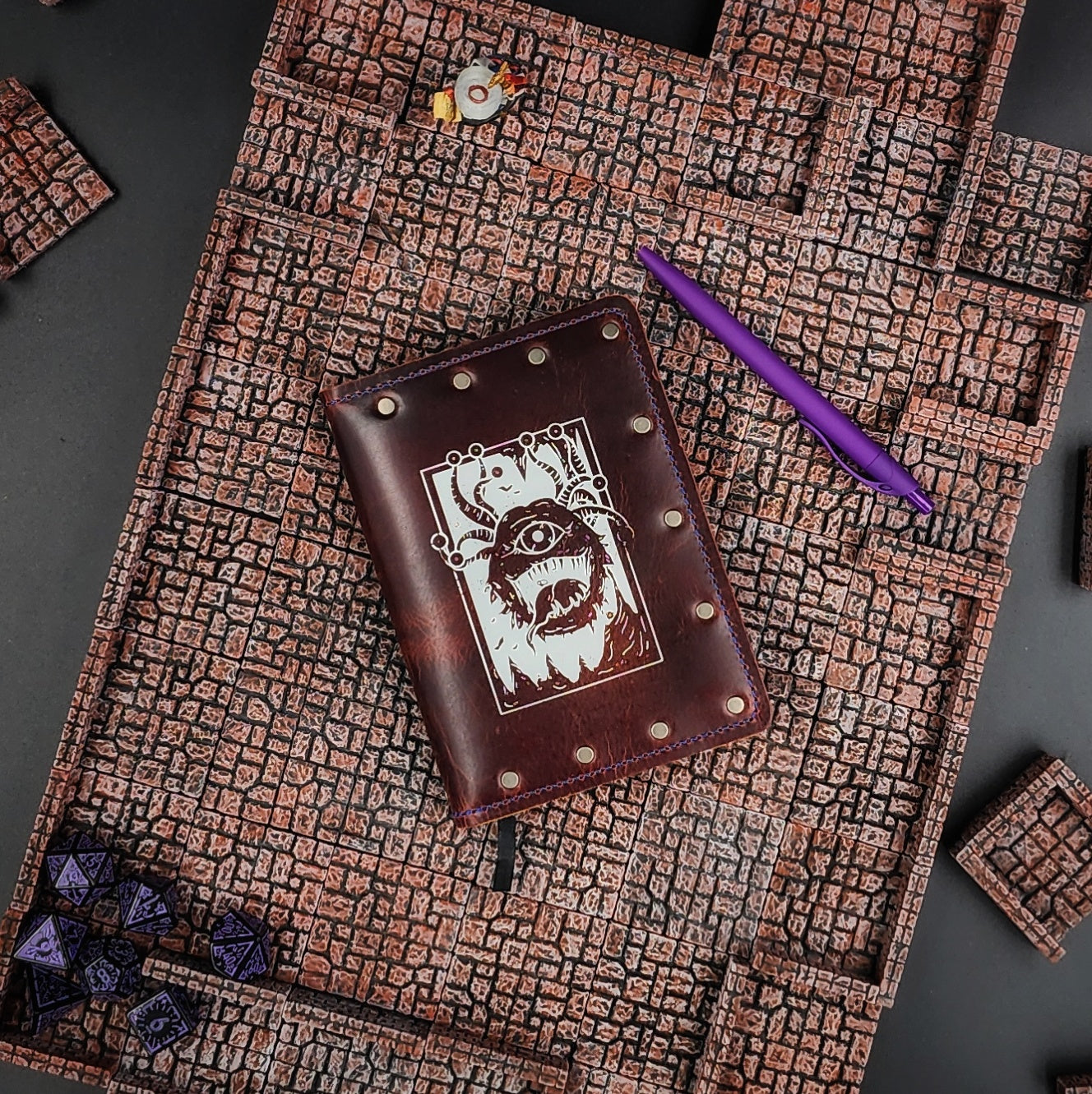 Handcrafted leather pocket adventure journal with hand-gilded Beholder design, shown on a tabletop game shop retail counter.