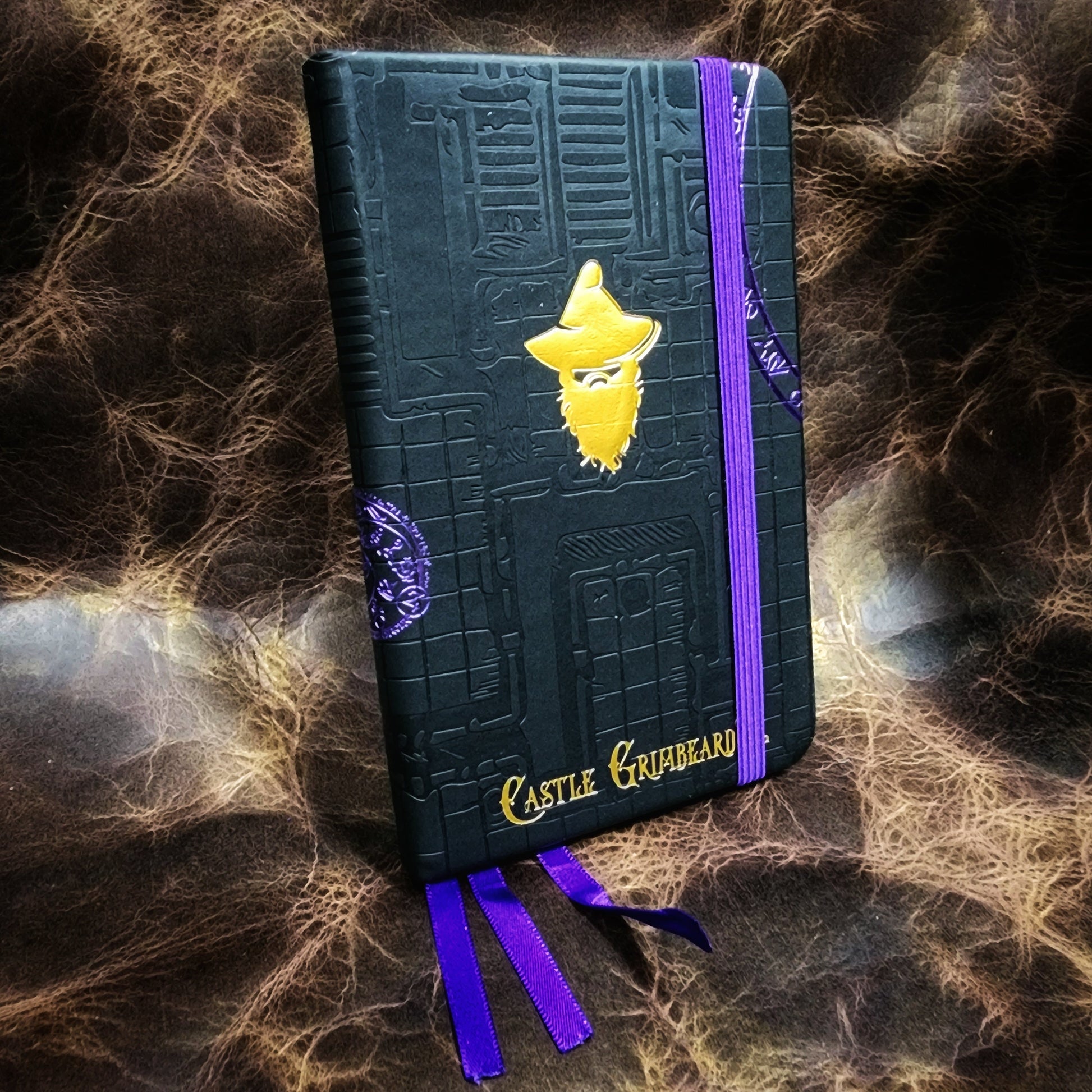 Hardcover black A6 pocket adventure journal refill insert featuring an embossed old school dungeon grid and gilded purple foil rune circles by Grimbeard Leather.