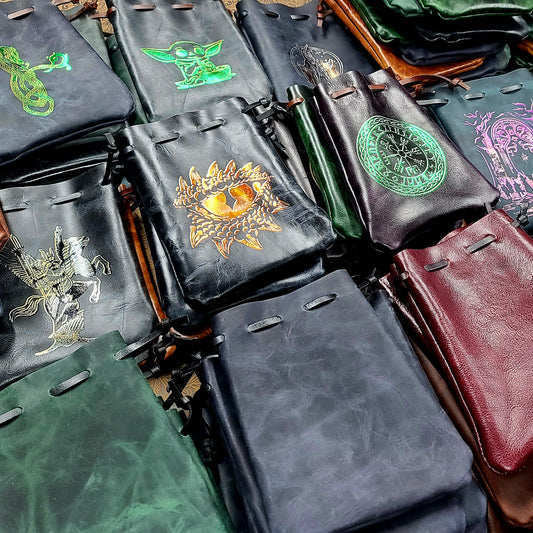 Handcrafted patterned and stamped leather dice bags in earthtones, blacks, grays, reds, blues, greens, purples, pinks, yellows, and golds for D&D players and retail store counters.