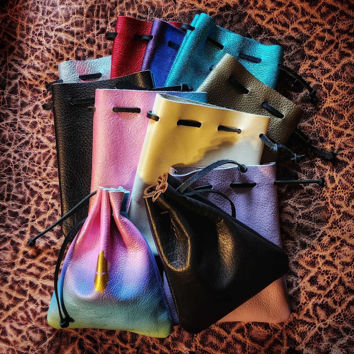 Handcrafted patterned and stamped leather dice bags in earthtones, blacks, grays, reds, blues, greens, purples, pinks, yellows, and golds for D&D players and retail store counters.