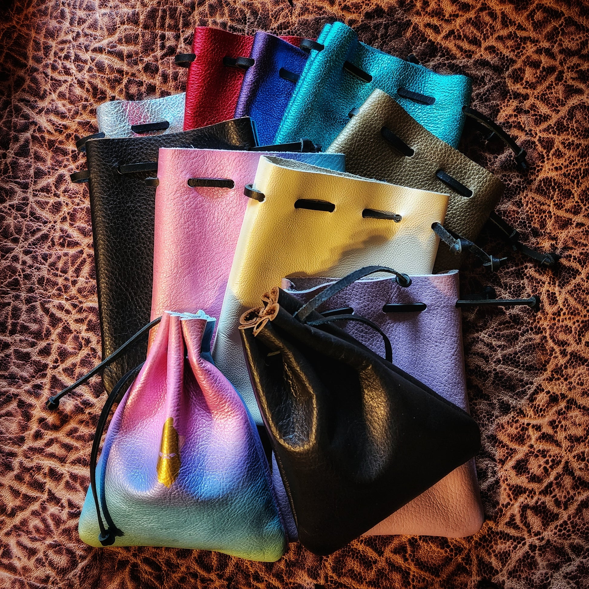 Handcrafted patterned and stamped leather dice bags in earthtones, blacks, grays, reds, blues, greens, purples, pinks, yellows, and golds for D&D players and retail store counters.